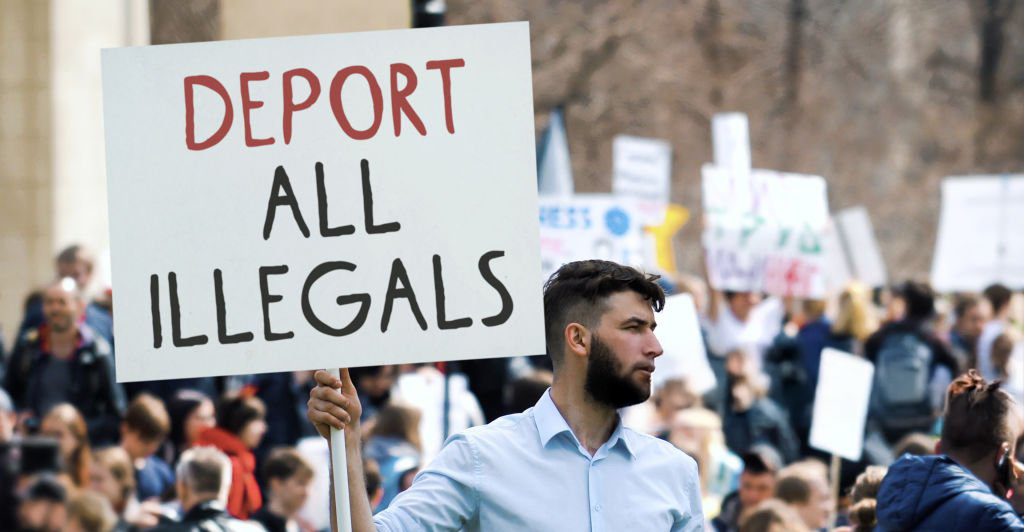 Protesters holding signs at London Protest 2025 rally led by Tommy Robinson, with banners about immigration and identity issues in the UK