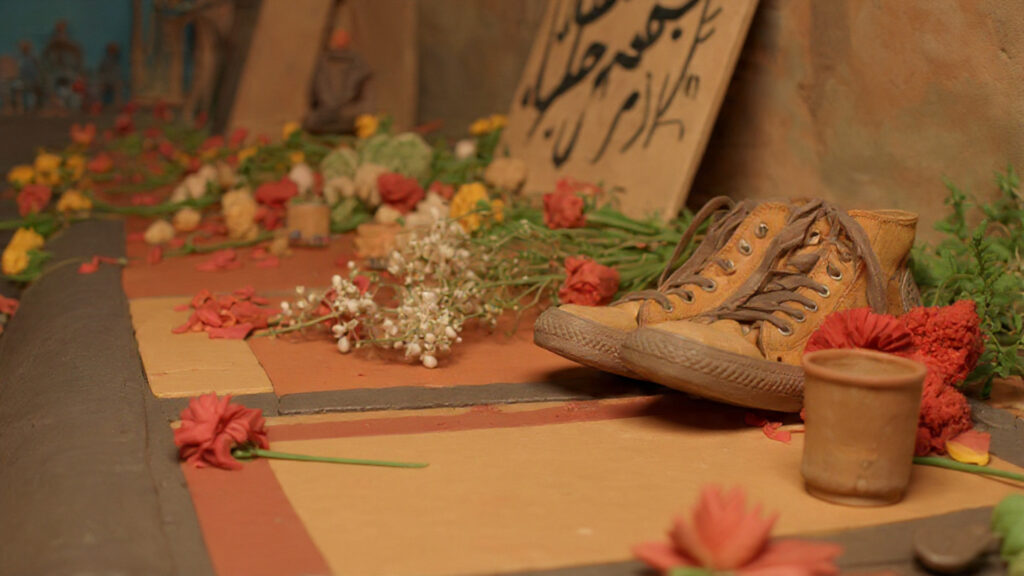 A makeshift street memorial with candles, flowers, and worn shoes on a Tehran sidewalk.
